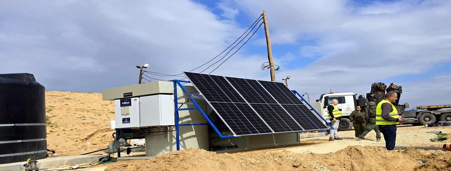יחידת טיפול של INFRABOX, המערך הסולארי במסגרת הכחולה ומשטח הבטון באתר מדברי מיושר, עם קווי מתח עיליים מאחור.