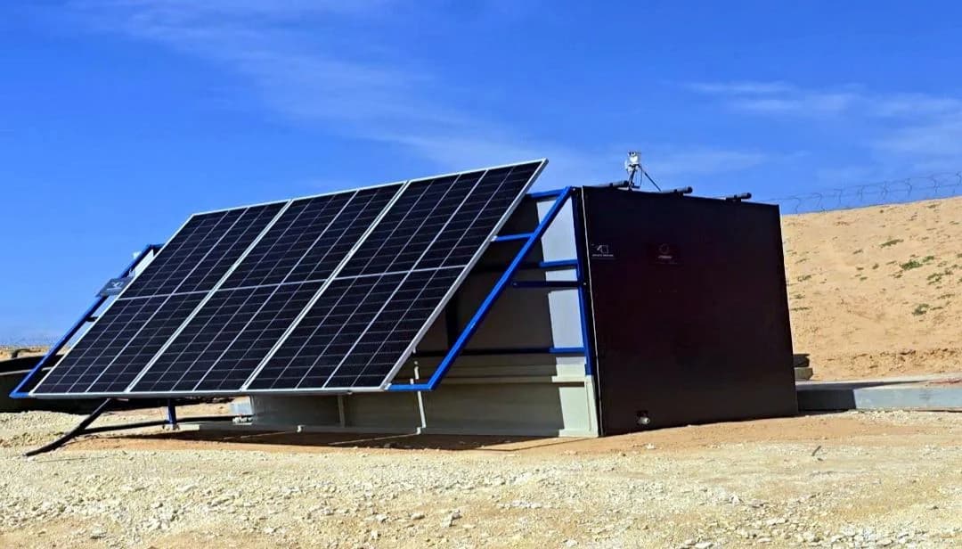 Un ensemble de panneaux solaires à cadres bleus incliné à côté d’une unité de traitement INFRABOX noire, sur une dalle dans le désert.