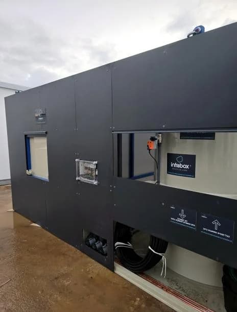 The rear service face of an INFRABOX unit with its access hatch open and bundled conduits running out to the pad.
