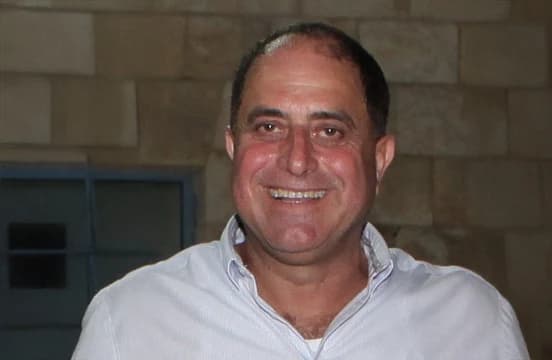 In a pale blue shirt, photographed in front of a stone wall and a barred window.