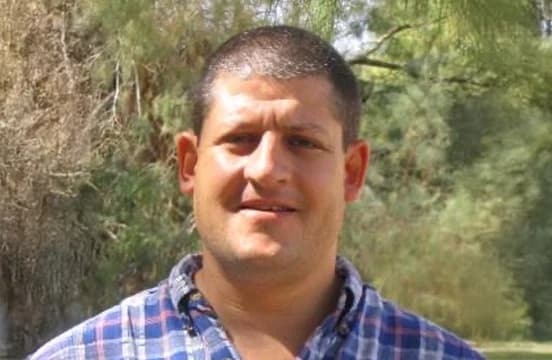 In a blue and red checked shirt, photographed outdoors with trees behind.