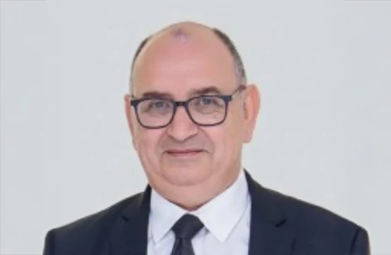 In heavy-framed glasses, a dark suit, a white shirt and a dark tie, photographed against a plain pale studio ground.