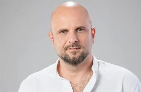 Shaven-headed and with a short beard, in an open-necked white shirt against a plain gray ground.