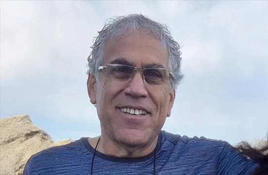 Gray-haired, in rimless glasses and a blue ribbed top, photographed outdoors against pale rock and open sky.